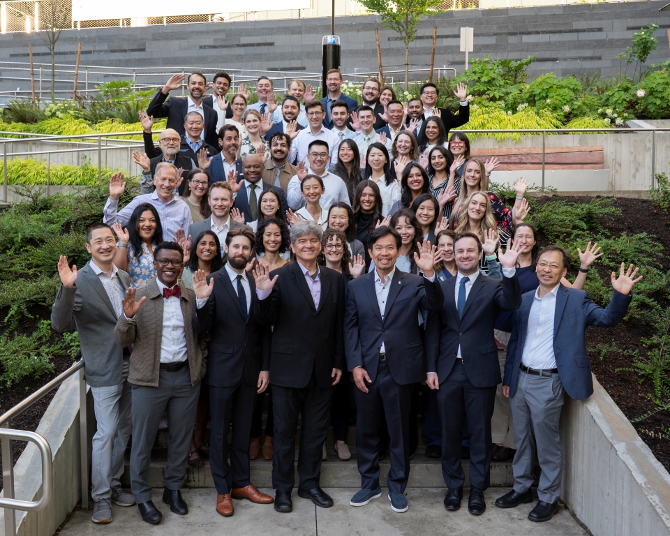 A large group of people dressed professionally pose smiling and waving at the camera.