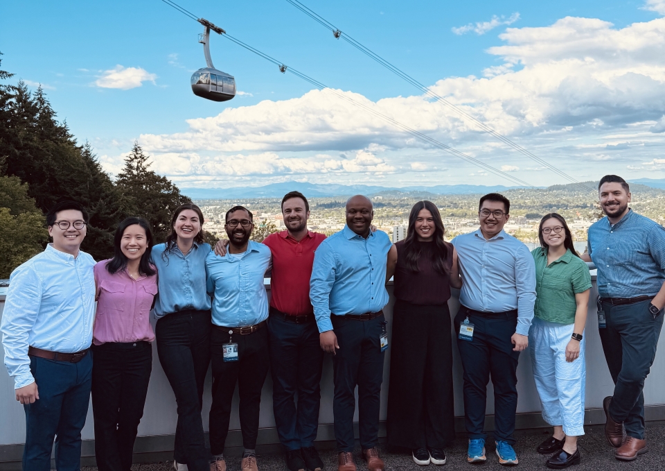 A group of 10 young people stand against a railing with the Portland Aerial Tram traveling behind them in a clear blue sky.