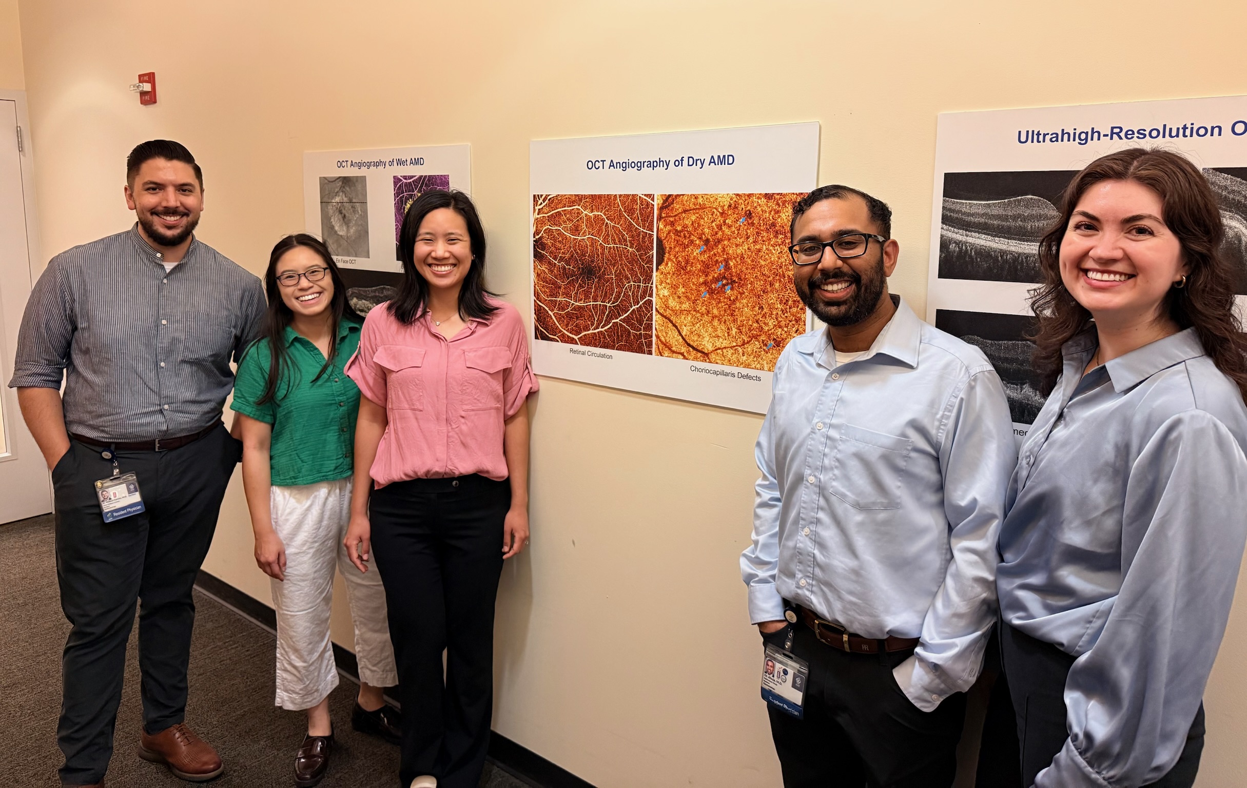 A group of five people pose together in front of a wall of OCT images published in academic journals.