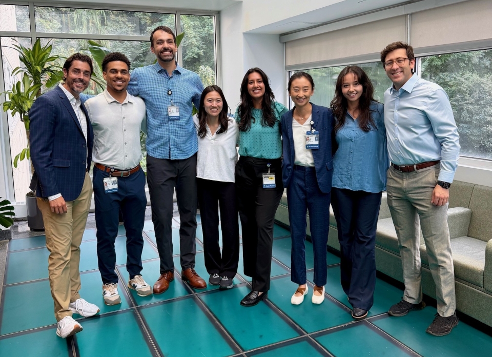 A group of people stand arm-in-arm smiling at the camera while standing on a green floor.