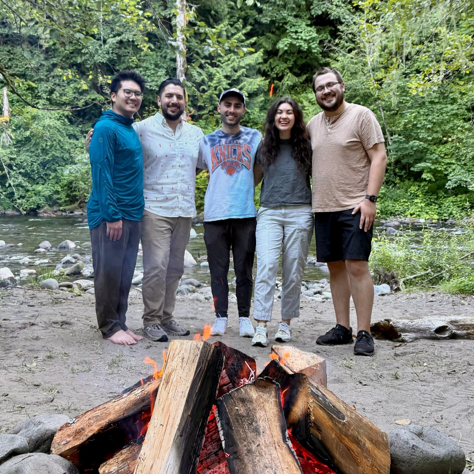 Group of five young people stand together in the woods in front of an open firepit