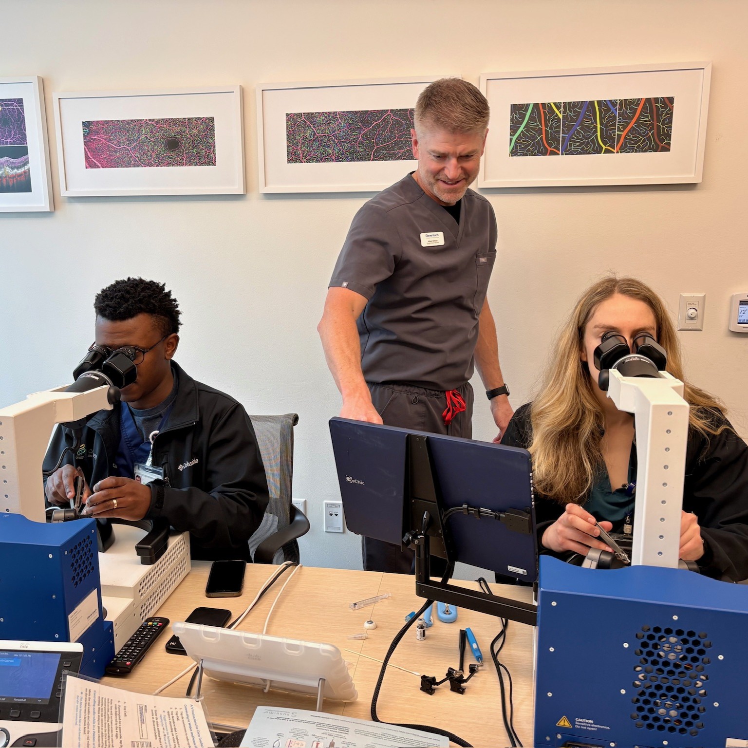 Two people sit looking through microscopes while a man stands between them to assist.