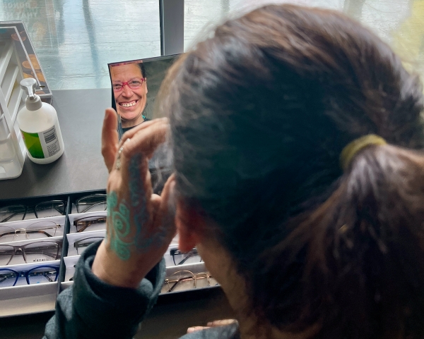 The camera looks over the shoulder of a women looking at her reflection in a mirror while she holds up a pair of glasses and smiles. 
