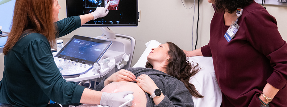 A pregnant person lies on an exam bed between two OHSU healthcare providers. All three are looking at a screen showing images from a scan.