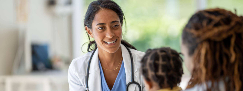 A young doctor holds a digital tablet while talking to a young child in her mother’s lap in a medical exam room.