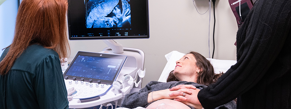 A pregnant patient looks at an image on a screen while a healthcare professional stands next to her, holding her hand. Another healthcare professional sits in front of the screen.