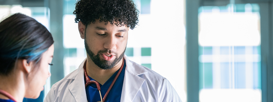 Doctor stands in a hospital near windows consulting with a healthcare worker who is beside him.