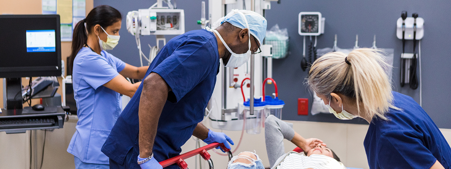 Two doctors wearing scrubs and masks examine a patient in the ER as a nurse prepares an IV.