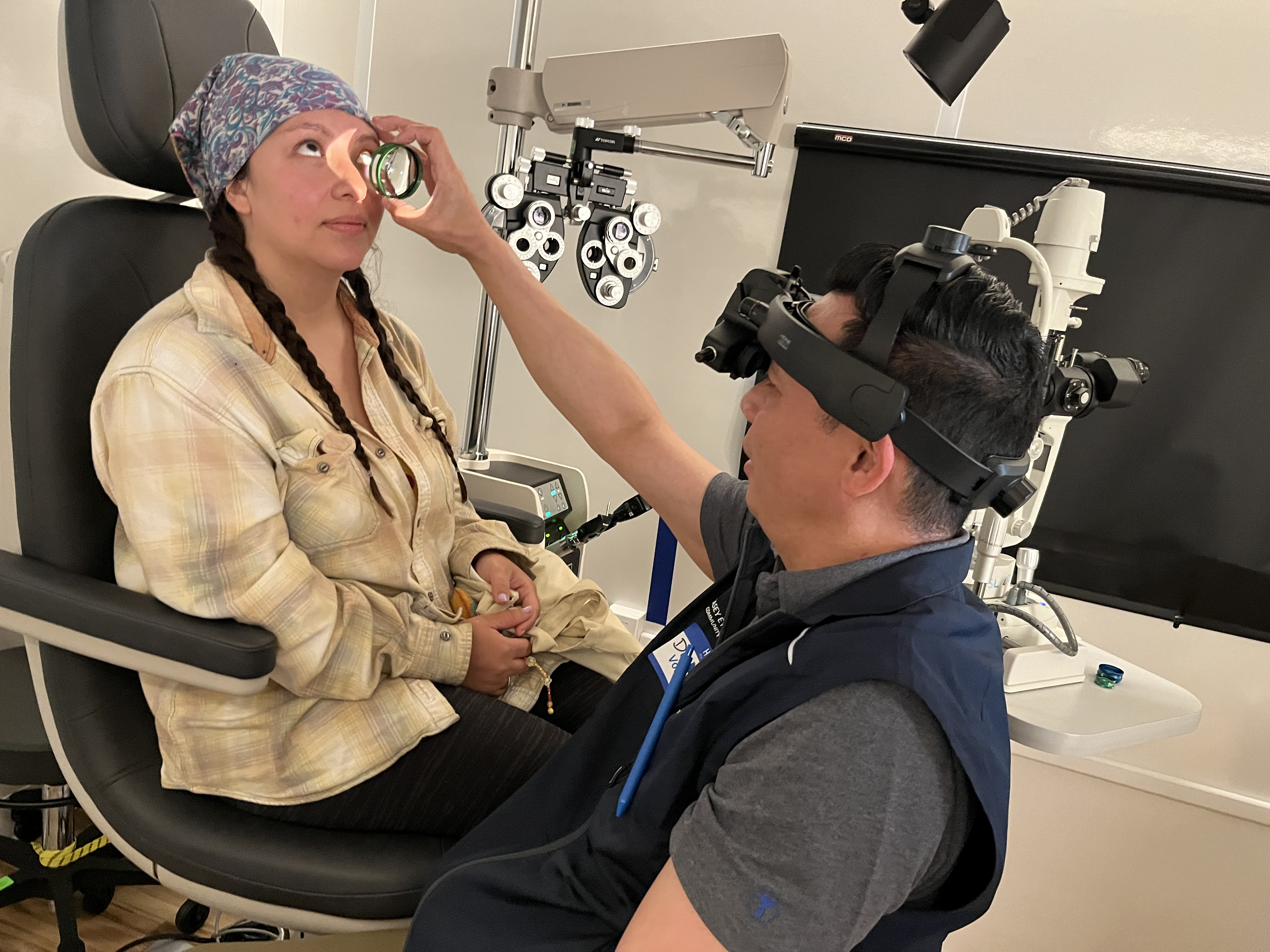 A doctor sits in front of a patient in an exam chair during an eye exam. He holds up a lens close to her eye while checking her vision.