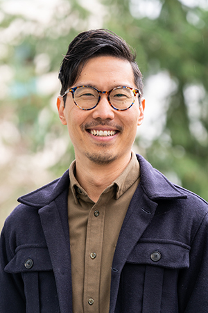 Professional headshot of man wearing glasses smiling at camera with natural background