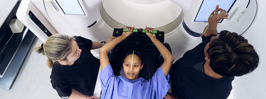 Patient positioned on a radiation therapy treatment table while two clinicians align laser guides before treatment.