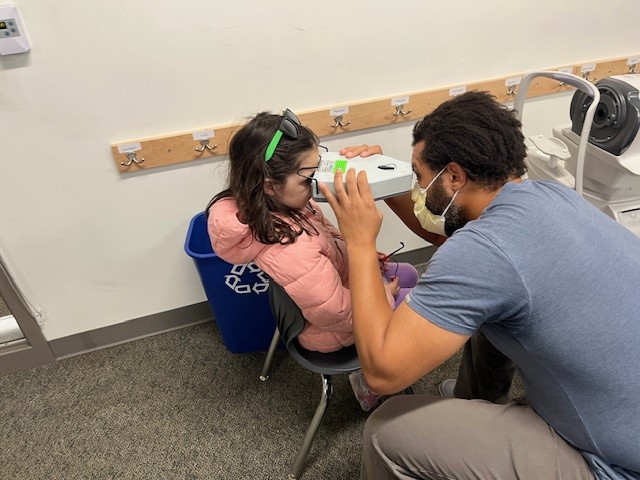 A man bends down in front of a small girl so she can look into a device that will screen her vision.