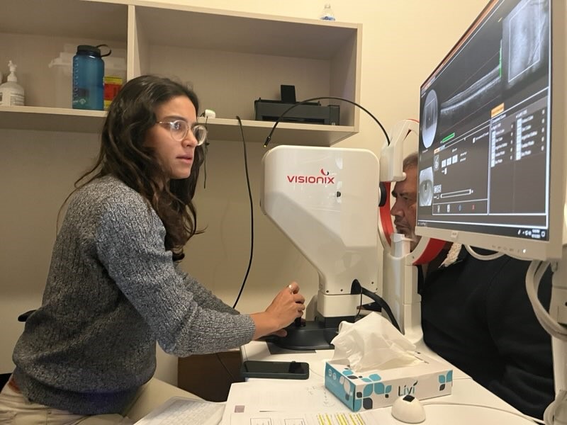 A woman operates a machine that takes pictures of the eye while a patient leans forward looking into the device.
