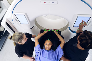 Patient positioned on a radiation therapy treatment table while two clinicians align laser guides before treatment.