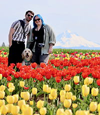 A man and a woman stand smiling, with their dog sitting in between them, in a flower field on a sunny day with a mountain visible in the background.