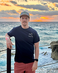 A man standing in front of wire fencing with the ocean behind him at sunset.