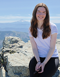 A woman sitting on a rock ledge of a mountain on a sunny day, with another mountain visible far off in the distance.