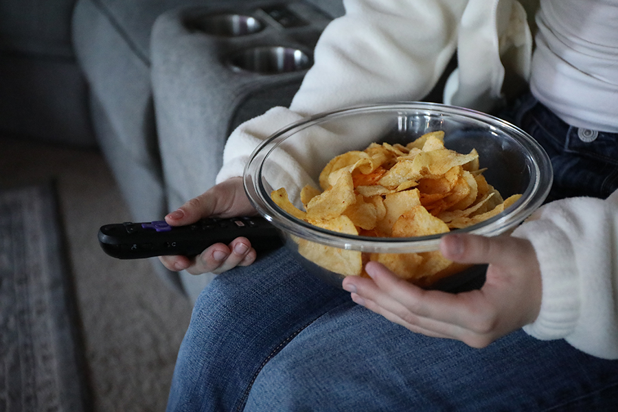 A person holds a bowl of potato chips in one hand and a TV remote in the other.