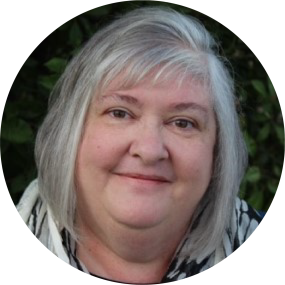 Rhonda Eppelsheimer, a person with white and gray hair wears a black and white scarf and smiles into the camera with greenery in the background.