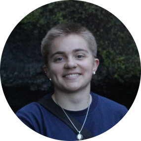 UCEDD Student Worker Nikolai D. Zylman, a person with short dark blond hair and olive skin, wearing a navy blue crew-neck t-shirt and a silver pennant necklace.