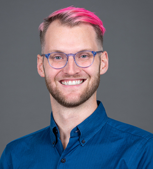Blair Peters, with pink hair, wearing a blue button-down shirt and blue glasses, against a grey background